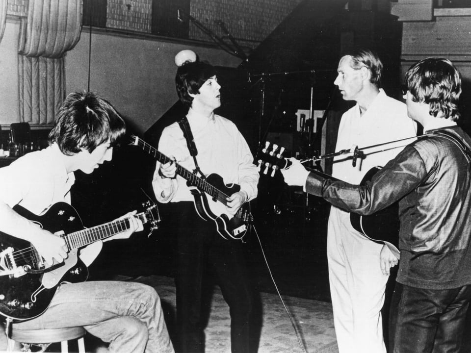 Drei Musiker mit Gitarre stehen um George Martin herum.