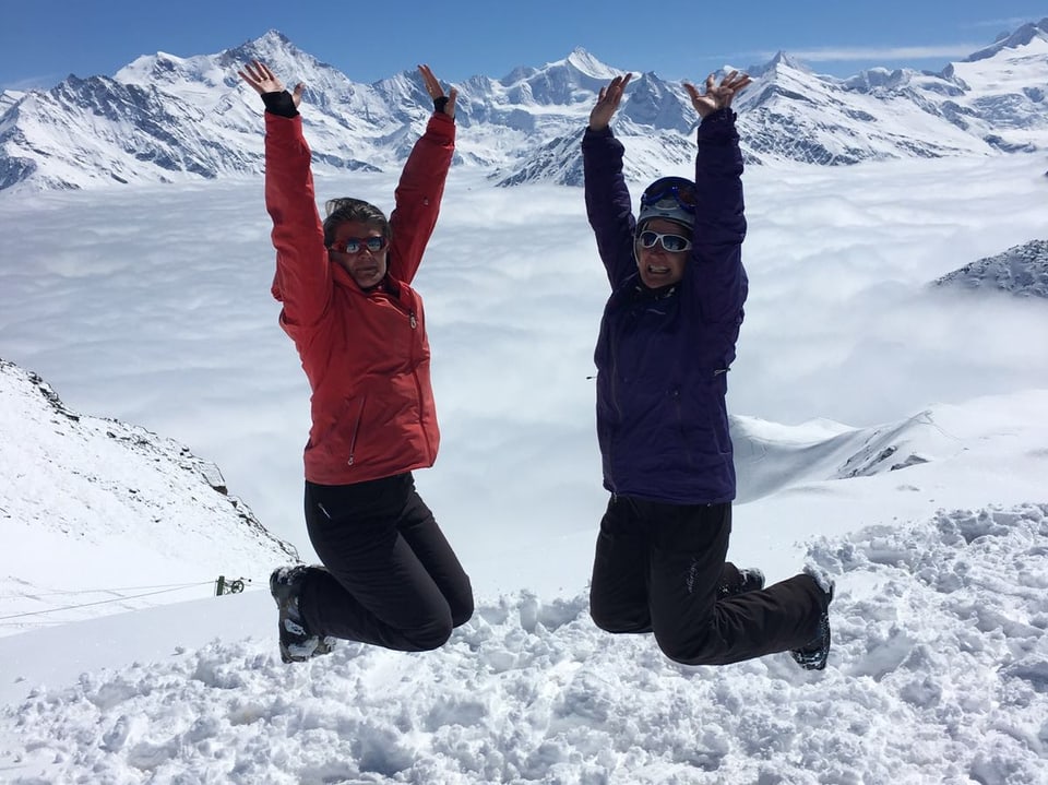 Zwei Frauen ins Skibekleidung und mit Skischuhen an den Füssen machen gleichzeitig einen Luftsprung. Im Hintergrund das Nebelmeer und darüber verschneite Berggipfel und einen blauen Himmel.