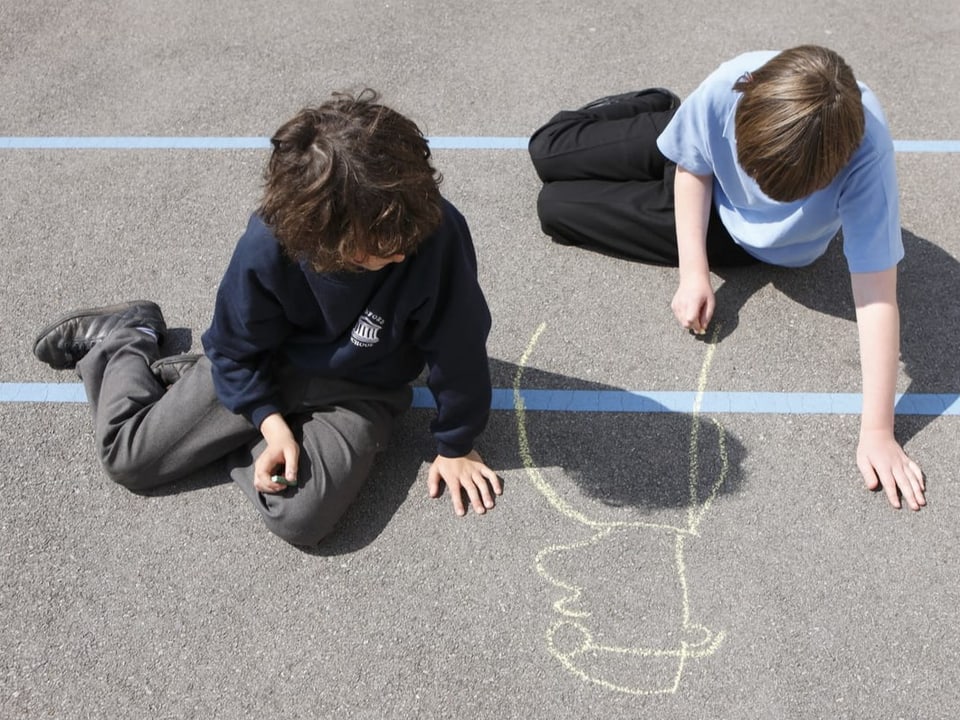 Zwei aus der Vogelperspektive fotografierte Kinder zeichnen mit Kreide auf einem Pausenplatz