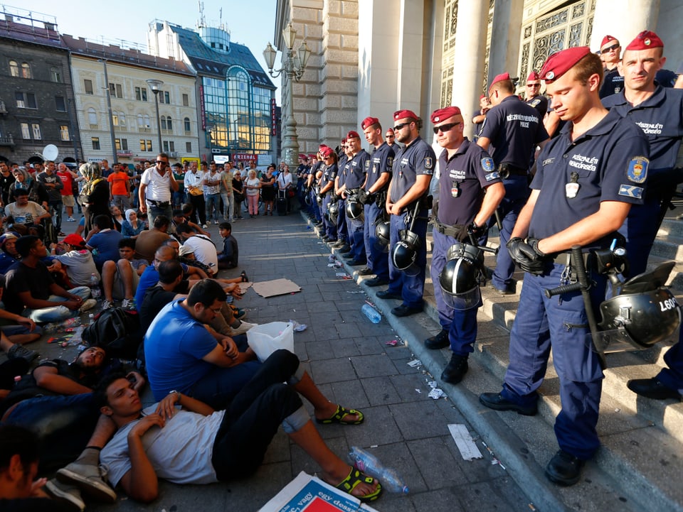 Links junge Männer, zum Teil am Boden, rechts zwei Reihen Polizisten mit rotem Beret, die den Bahnhof versperren.