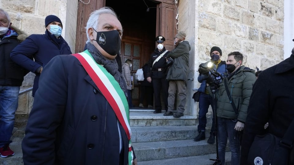 Mann mit Maske und Schärpe in den Farben der Tricolore steht vor einer Kirche. Medienleute daneben.