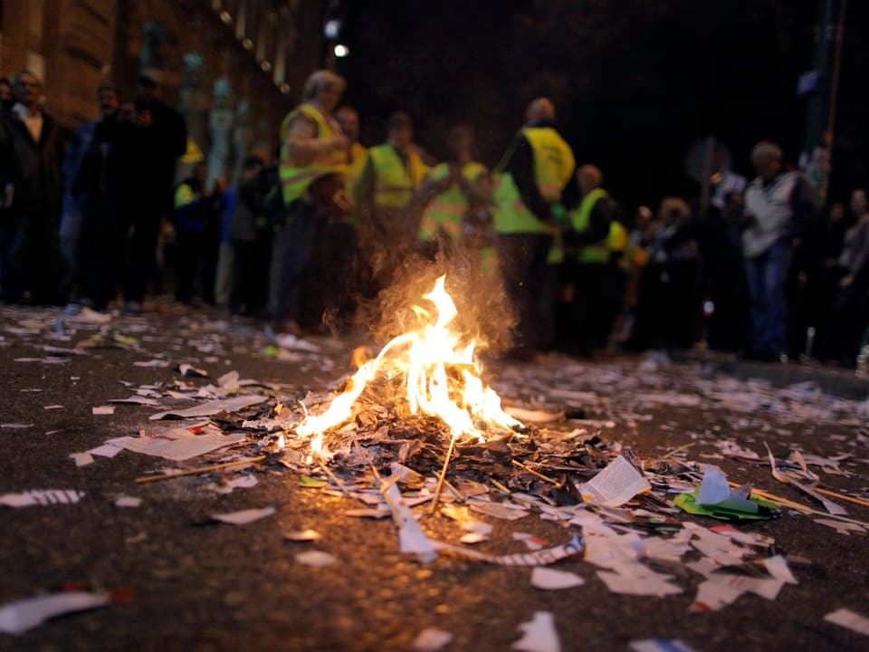 Ein kleiner Haufen brennender Abfall bei Nacht, im Hintergrund Demonstranten.