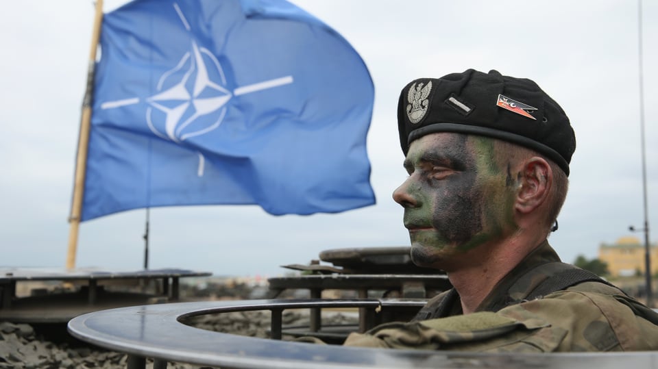 Tarngeschminkter Soldat mit Barrett blickt aus Geschützstand, dahinter eine im Wind flatternde Nato-Flagge.