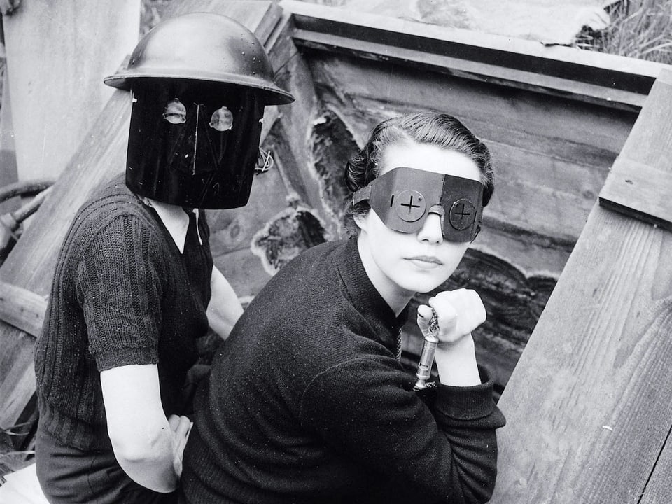 Zwei Frauen mit selbstgebastelten Maskensitzen nebeneinander und schauen über die Schulter in die Kamera.