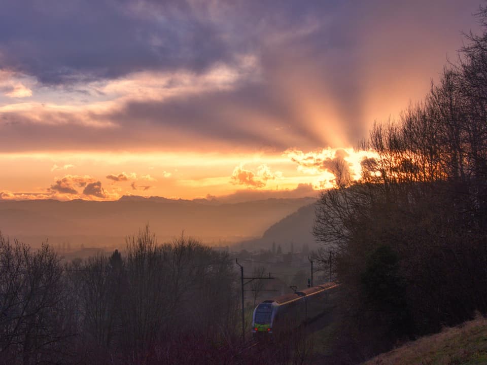 Blick ins Tal mit einem Zug im Vordergrund. Im Hintergrund färbt die aufgehende Sonne den Himmel über den Bergen gelblich.