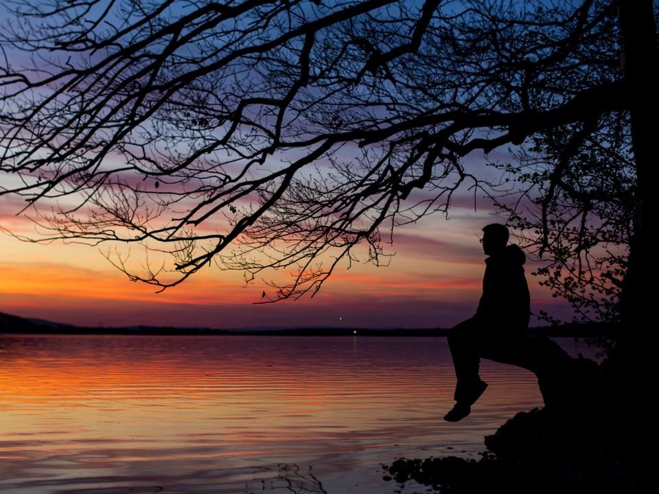 Dämmerung an einem See mit orangem bis violettem Himmel. Dieser spiegelt sich im Wasser. Am Ufer sitzt ein Mann unter einem Baum im Gegenlicht. 