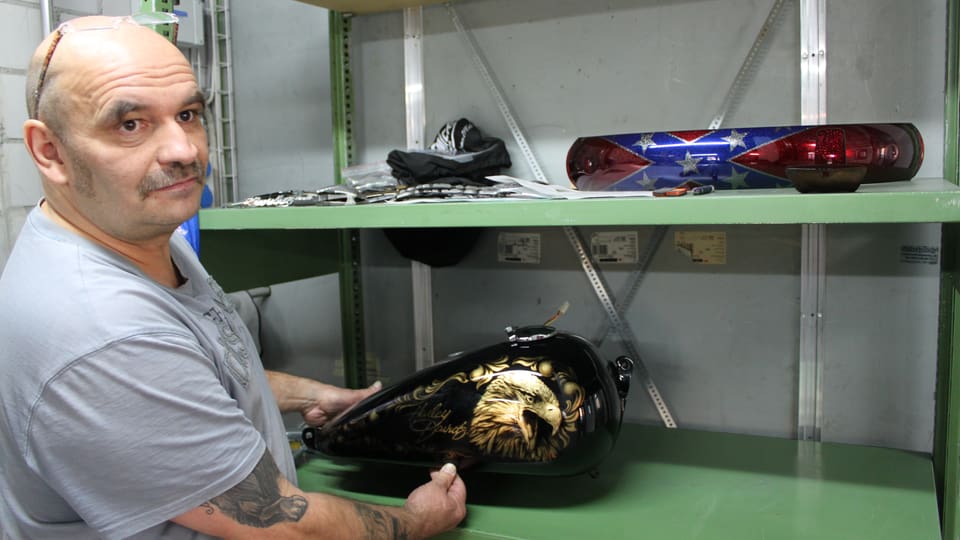 Mann aus der Harley-Werkstatt zeigt, was er gemacht hat. Ein Teil eines Töffs ist zu sehen. 