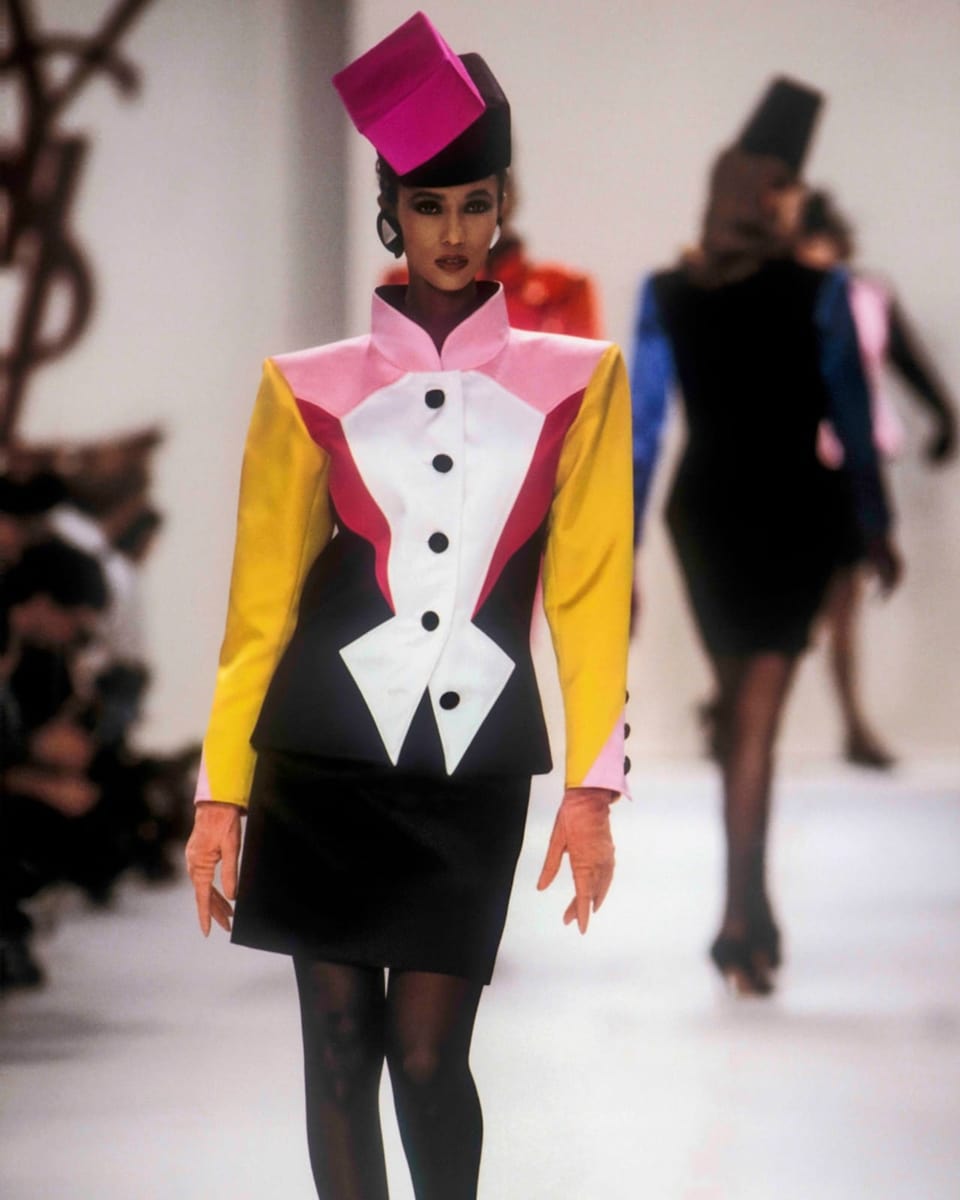Woman with a square, pink hat and top made of yellow, pink, red, white and black, square shapes, with a collar.