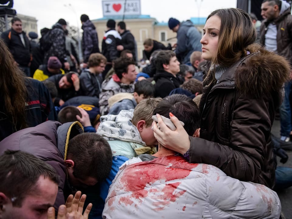 Ein Mann mit blutverschmierten Kleidern wird von einer jungen Frau umarmt, dahinter sind zahlreiche Menschen zu sehen.