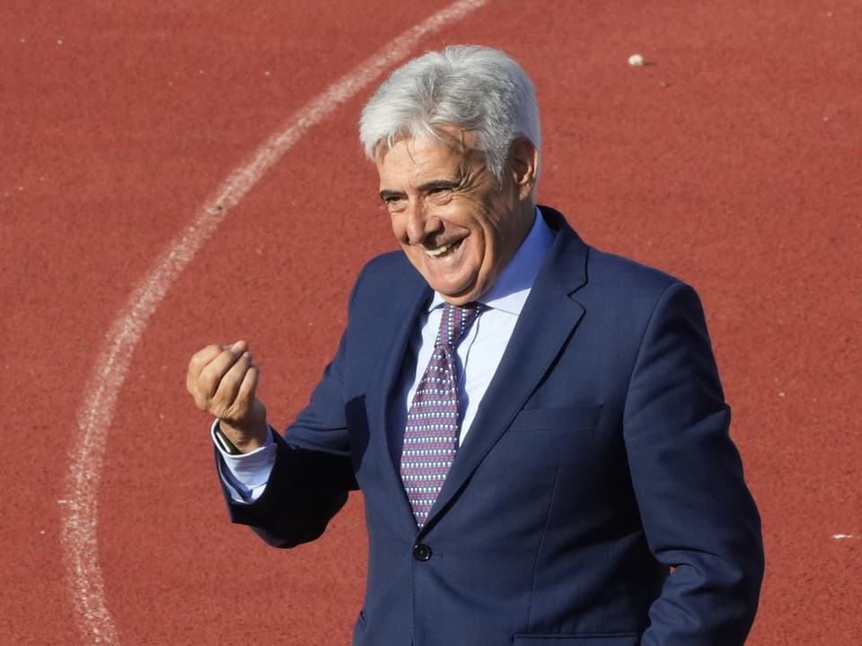 Älterer Mann in Anzug und mit grauen Haaren lächelt und winkt auf einer Sportbahn.
