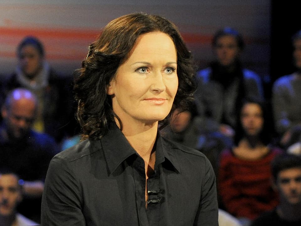 Eine Frau mit gelocktem dunklen Haar scheint ihrem Gesprächspartner in der TV-Show gespannt zuzuhören. Sie lächelt leicht und trägt eine schwarze Bluse. 