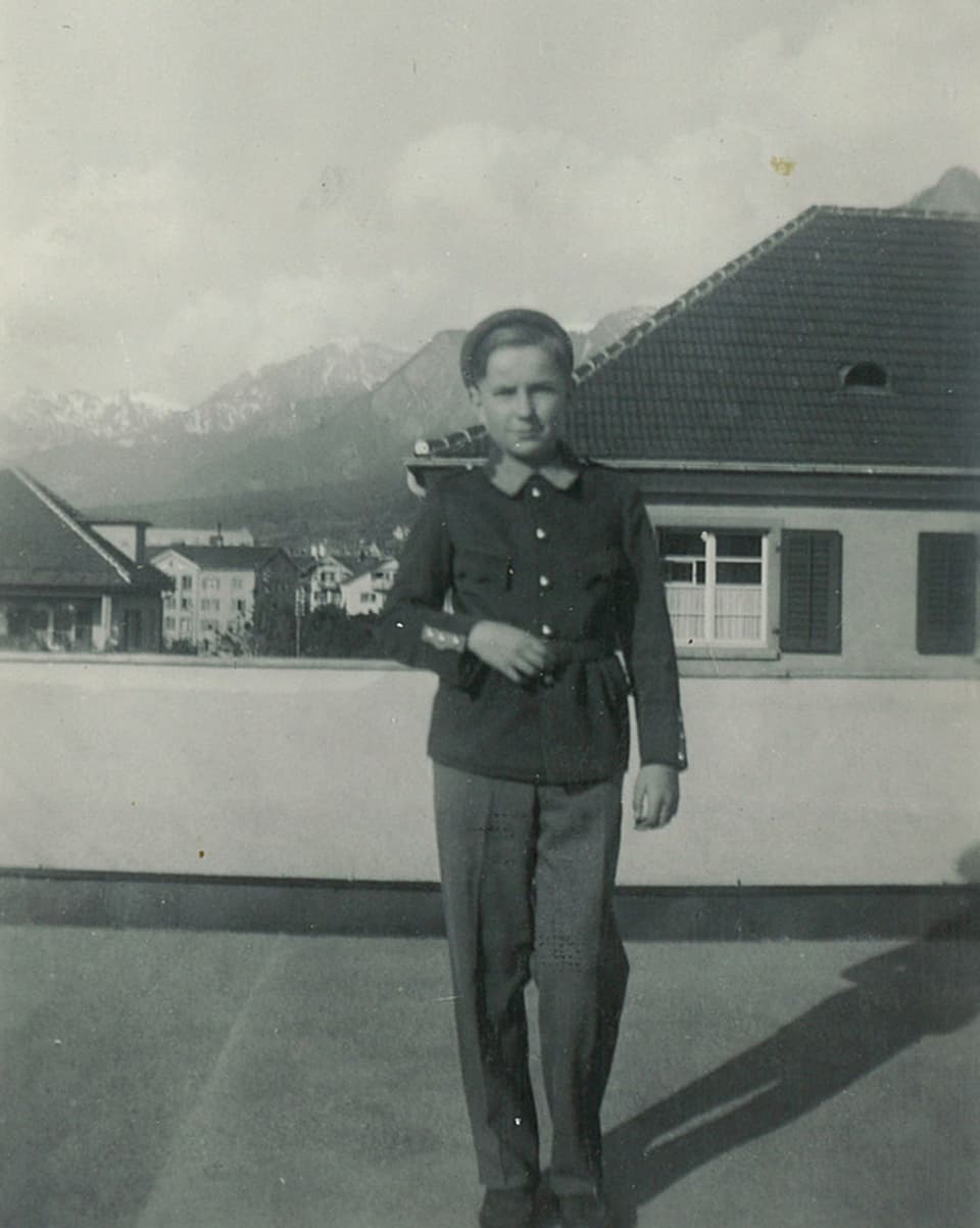 Der junge Bursche steht auf einer grossen Terrasse. Im Hintergrund sieht man Häuser und ein Alpenpanorama.
