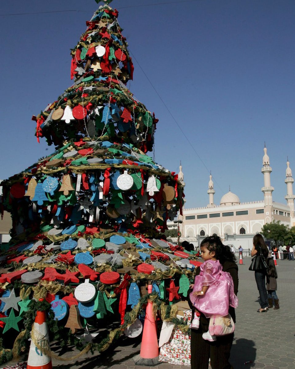 Ein bunter Weihnachtsbaum steht auf einem öffentlichen Platz in Dubai. Eine Frau mit einem Kind laufen daran vorbei.
