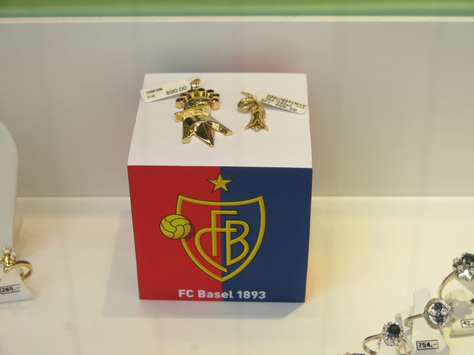 Podest für zwei kleine Schmuckstücke, dekoriert mit FCB-Logo. Darauf zwei goldene Baselstab-Anhänger. 