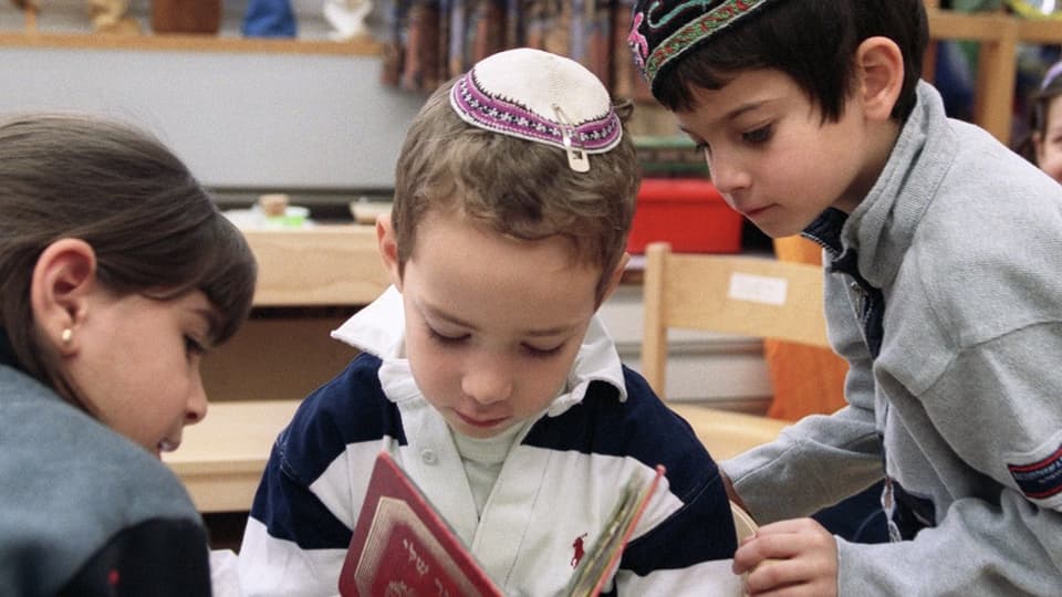 Ein Junge mit Kippa liest im jüdischen Kindergarten in der Bibel, zwei Kinder neben ihm schauen in das Buch