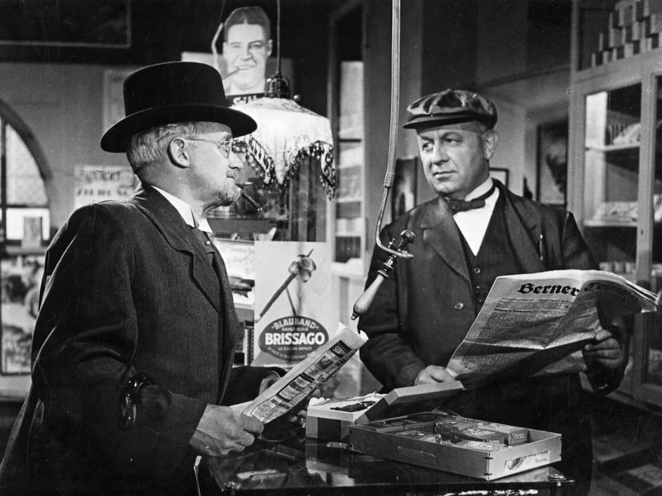 Zwei Männer stehen in einem Laden, beide mit einer Zeitung in der Hand. Sie schauen sich mit bestimmtem Blick an.
