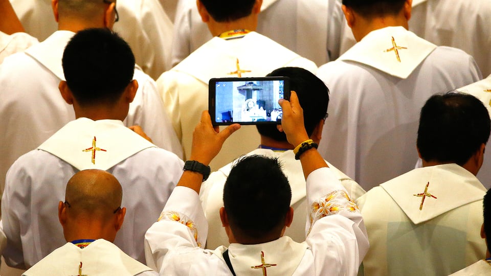Viele Priester von hinten. Einer hält ein Tablet hoch und filmt den Papst bei seiner ersten Ansprache in Manila.