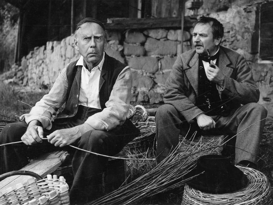Zwei Männer bei der Herstellung eines Besens aus Ruten. Der Mann zur Rechten raucht Pfeiffe und hat einen Schnauzbart.
