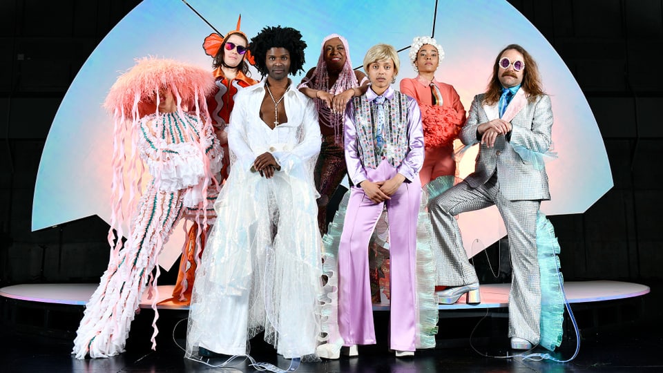 Auf einer Bühne stehen sieben Personen mit extravaganten Kostümen
