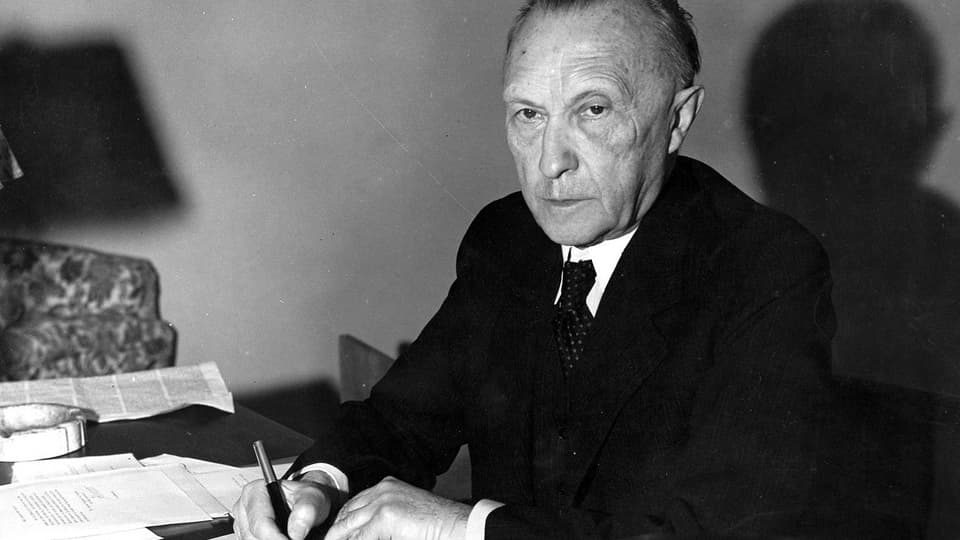 Konrad Adenauer, an einem Schreibstisch sitzend, mit Stift in der Hand, in die Kamera schauend.