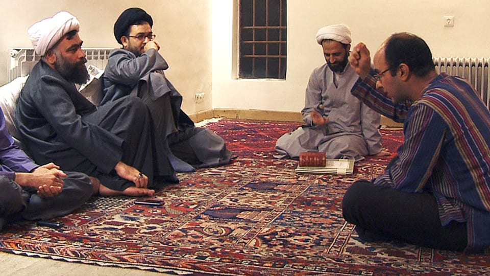 Vier Männer sitzen auf einem Teppich, drei tragen Turbane, einer gestikuliert.