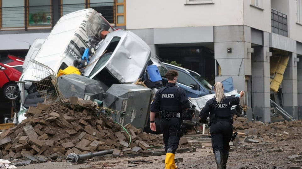 Polizist und Polizistin laufen auf der Strasse, im Hintergrund übereinandergestapelte Autos