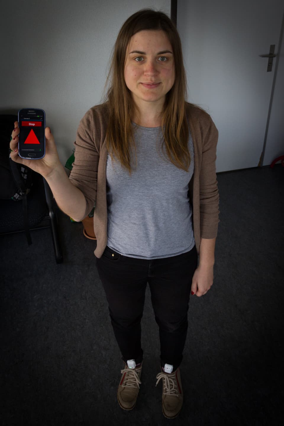 Eine junge Frau hält ein Smartphone in der Hand, an den Füssen sind Sensoren befestigt.