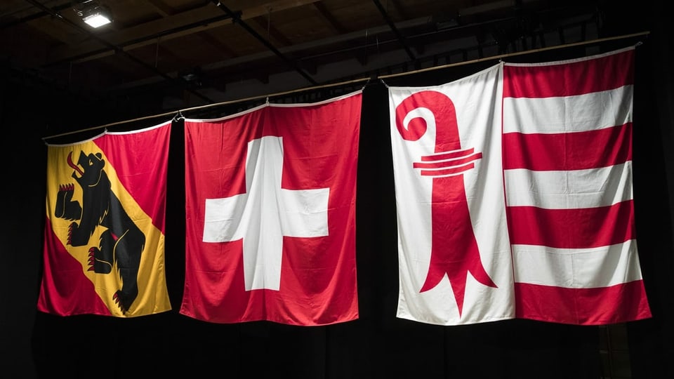 Drei Wappen-Fahnen hängen an einer Stange; Bern, die Schweizer Fahne und eine von Jura.