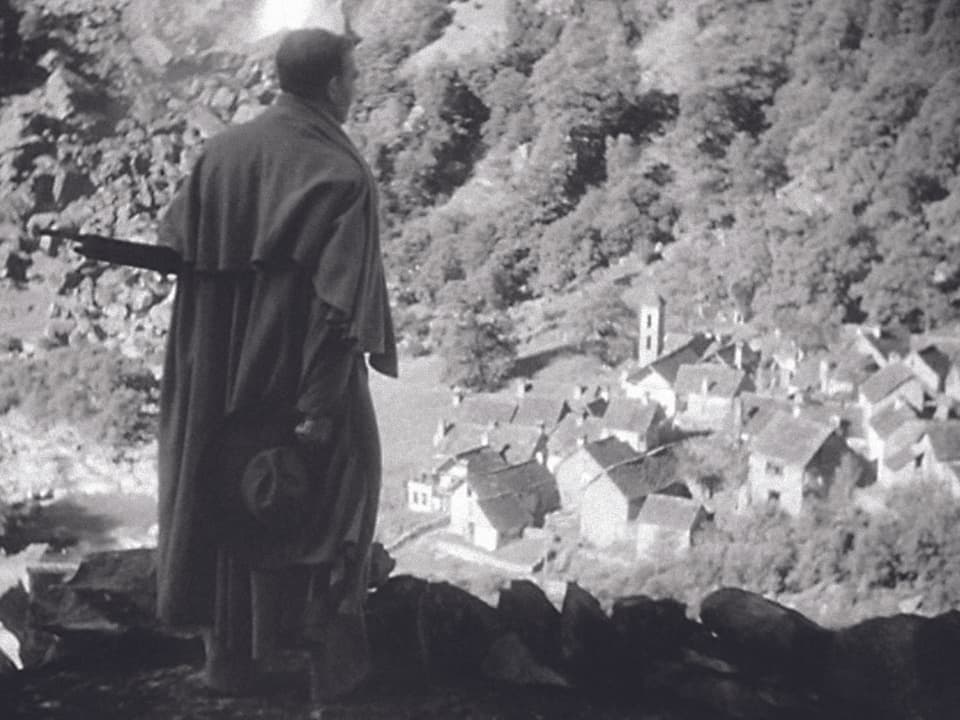 Filmszene in Schwarzweiss: EIn Man im Mantel schaut von einem BErg auf ein kleines Tessiner Dorf.