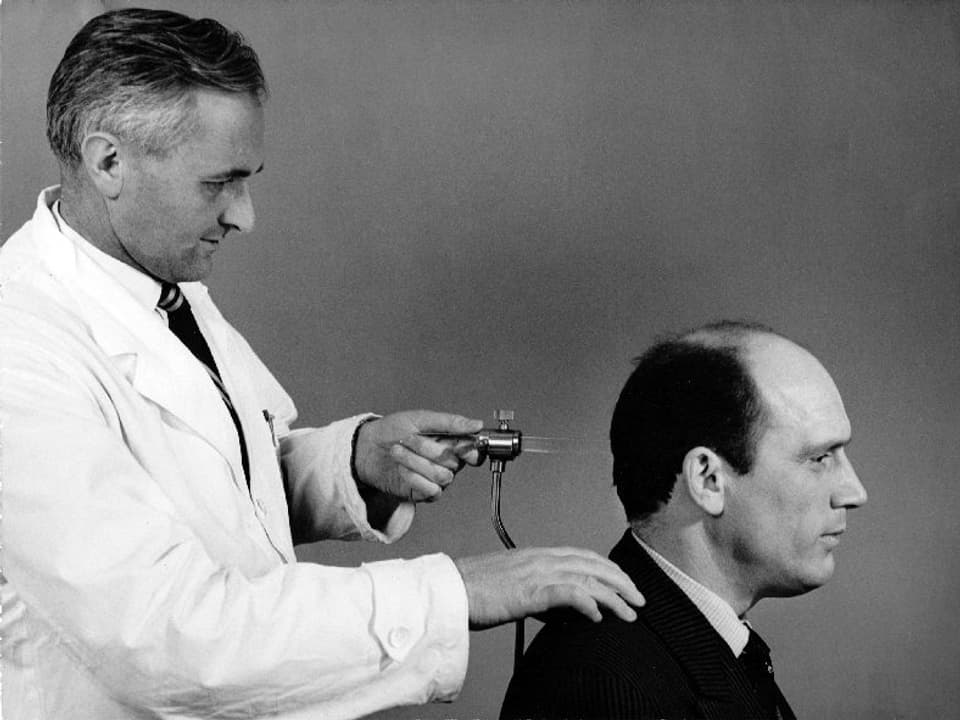 Ein Mann im Arztkittel hält eine Apparatur mit Glasrohr gegen den Kopf des vor ihm sitzenden Mannes.