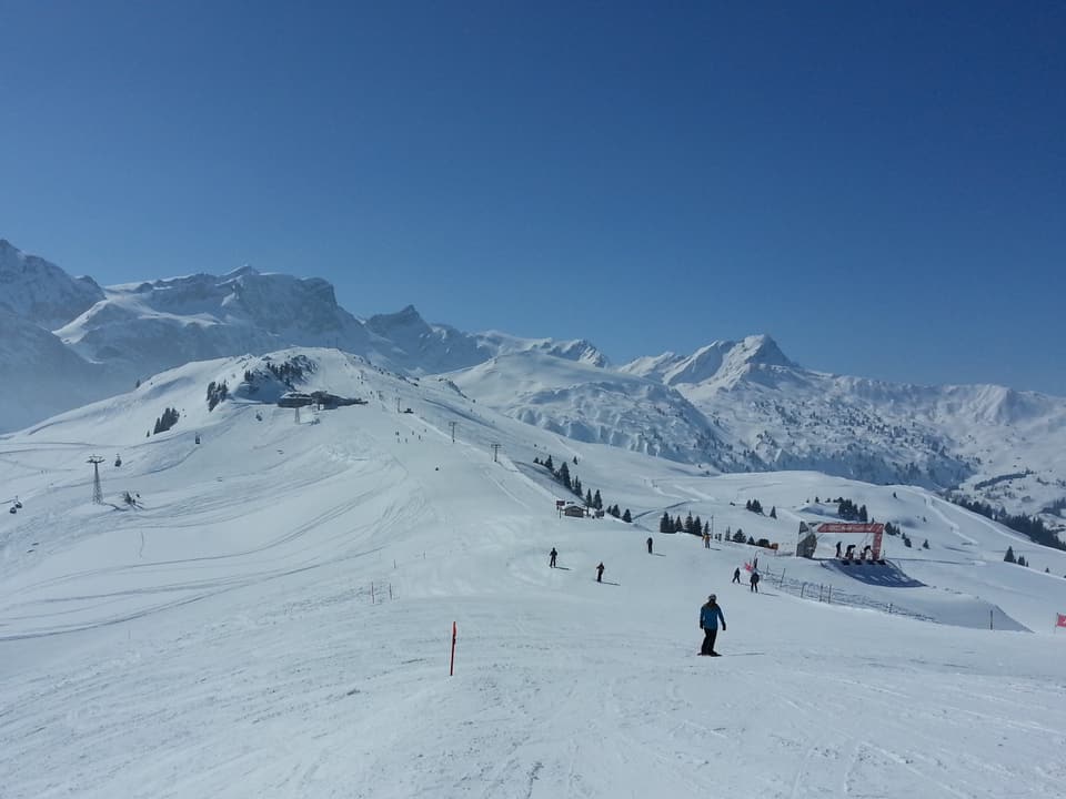 Skipisten, Skilifte, dahinter Berglandschaft mit Schnee. Darüber stahlblauer Himmel.