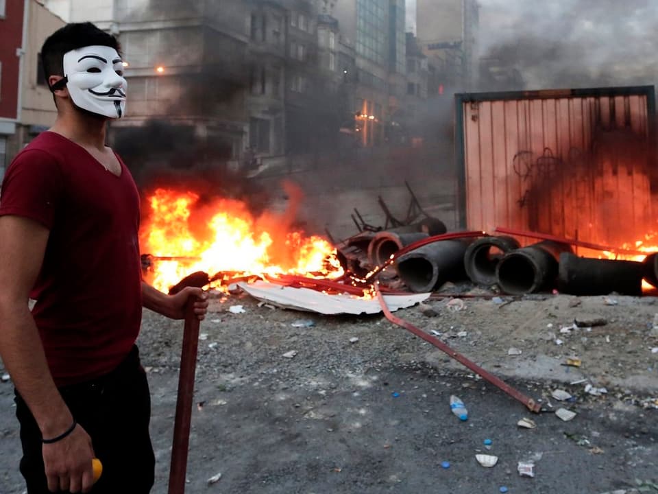 Maskierte Person vor brennender Barrikade.