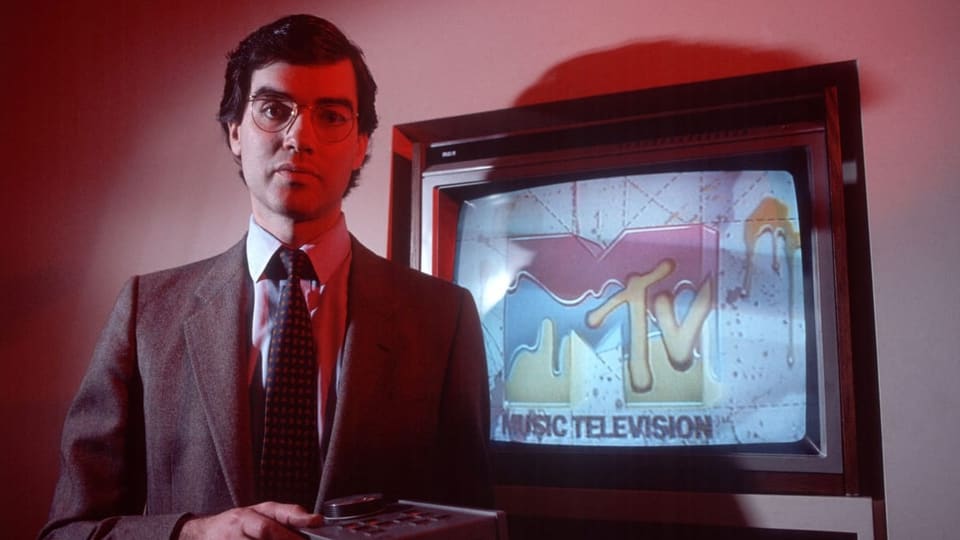 Ein Mann mit Anzug und Krawatte steht vor einem TV-Gerät, das das MTV-Logo zeigt.