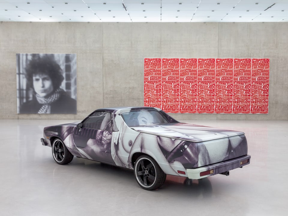 Auto mit Frauen bedruckt steht im Museum, an der Wand hängen ein Foto und ein farbiges Gemälde.