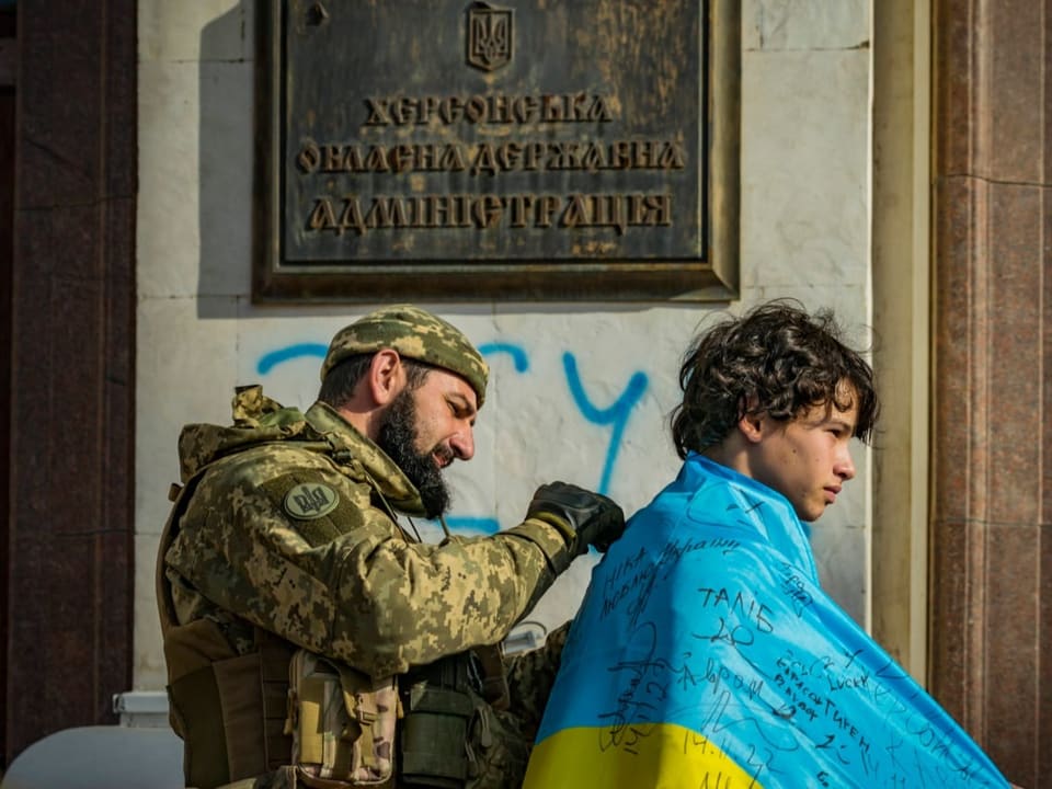 Ein Junge trägt eine Ukraineflagge über der Schulter. Ein Soldat steht hinter ihm und unterschreibt die Flagge.