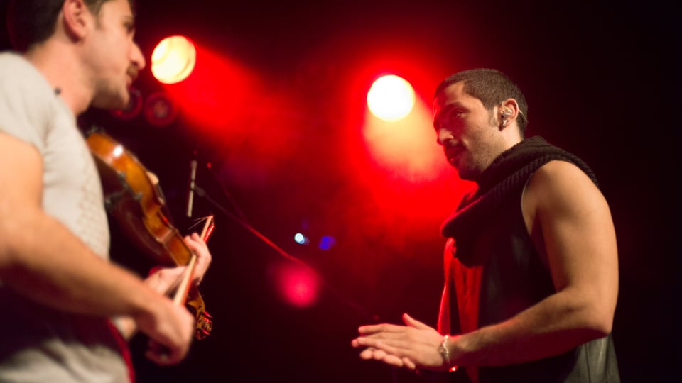 Ein Mann mit Geige und ein Mann, der klatscht, stehen auf einer rot beleuchteten Bühne und schauen sich an. 