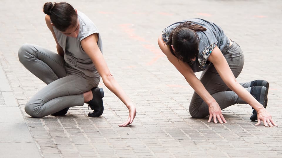 Zwei Frauen knien am Pflasterstein-Boden und führen eine Performance auf.