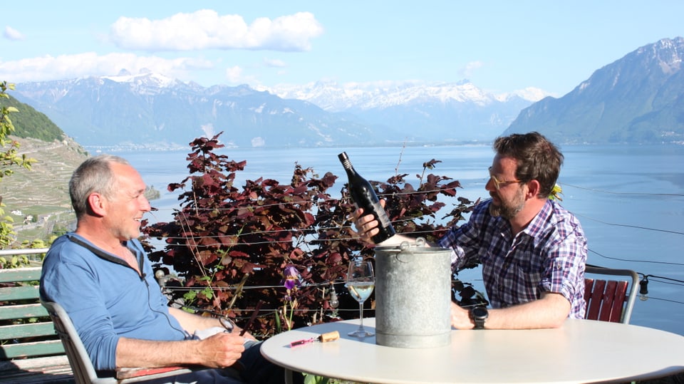 Nik Hartmann sitzt mit Weinbauer auf einer Terasse bei einem Glas Wein, im Hintergrund See und Berge. 