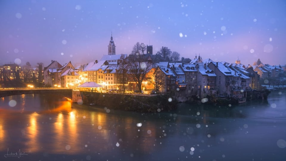 Stadt in der Dämmerung an einem Fluss mit Lichtern und einigen Schneeflocken. 
