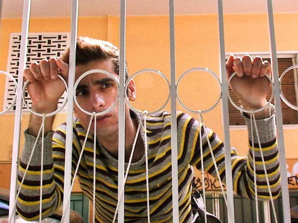 Filmstill: Ein junger Mann hinter dem Gitter eines Balkons.