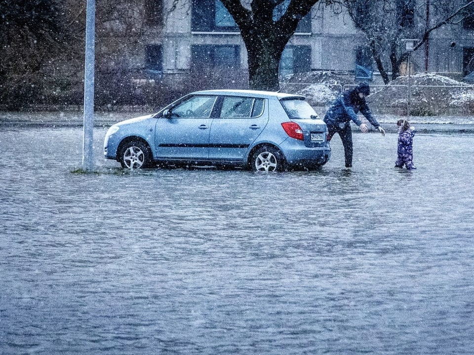 Ein Auto steht auf einem überfluteten Parkplatz. Zwei Personen stehen daneben.