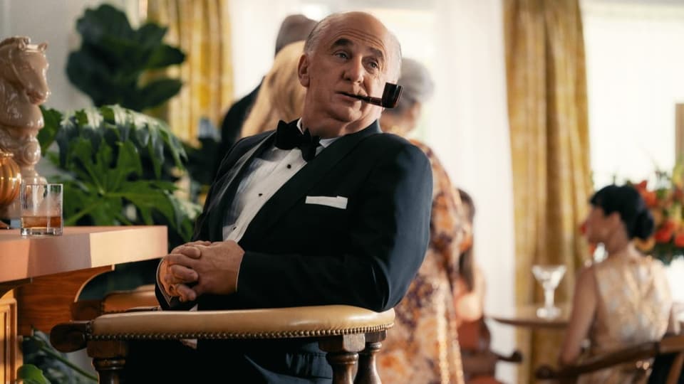 FIlmszene: Ein Mann mit Anzug und Pfeife im Mund sitzt an einer Bar.