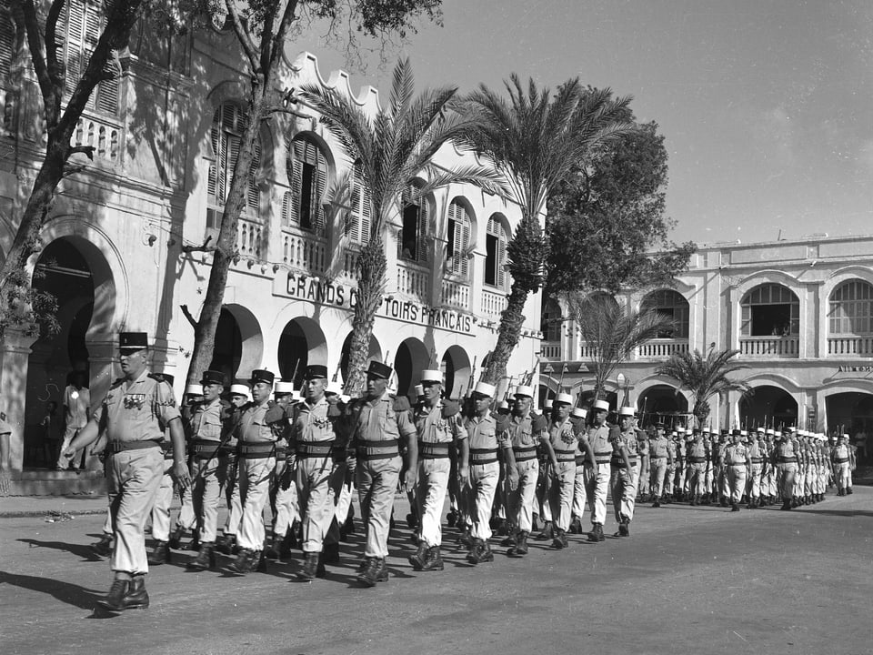 Soldaten Marschieren in Formation auf einem mit Palmen gesäumten Exerzierplatz.