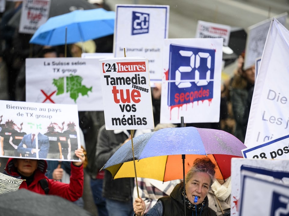 A woman holds a protest sign and rainbow umbrella during rain at the protest.