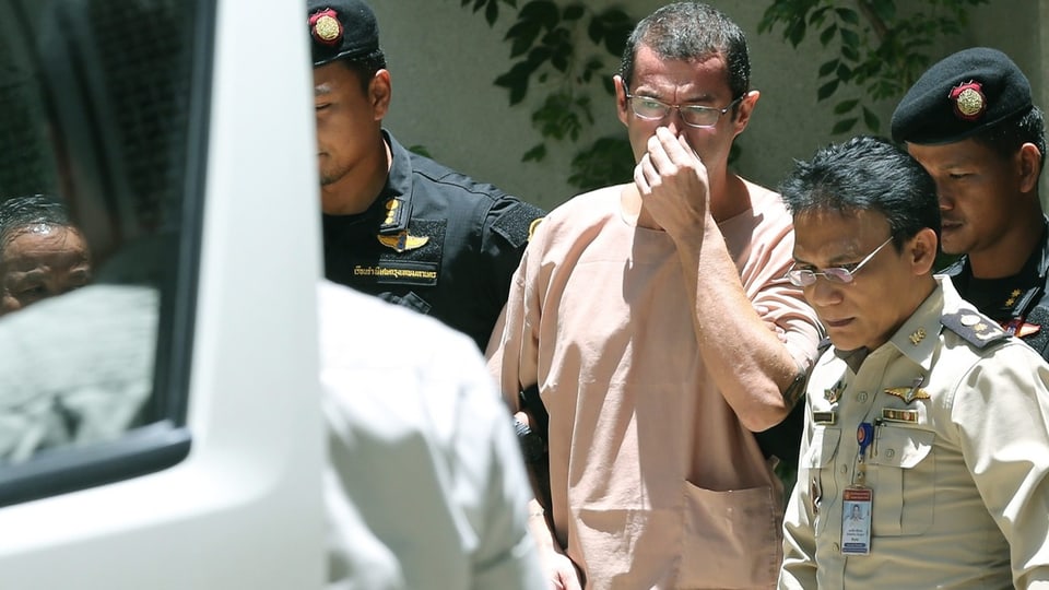 Mann mit kurzem Haar und Brille in lachsfarbenem T-Shirt wird von drei thailändischen Polizisten abgeführt.