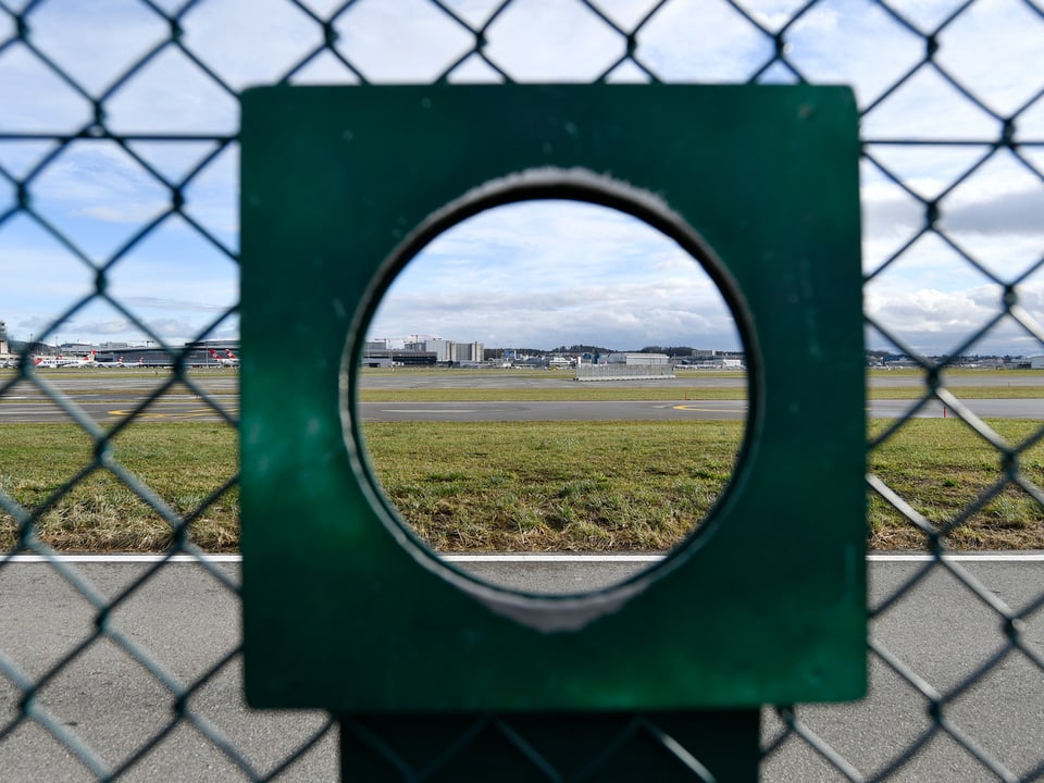 Zaun mit einem Loch. Dadurch hat man Blick auf das Flughafengelände.