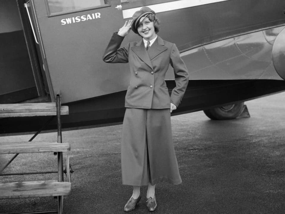 Schwarzweissbild, eine Frau mit langem Rock und Hut salutiert vor einem kleinen Flugzeug