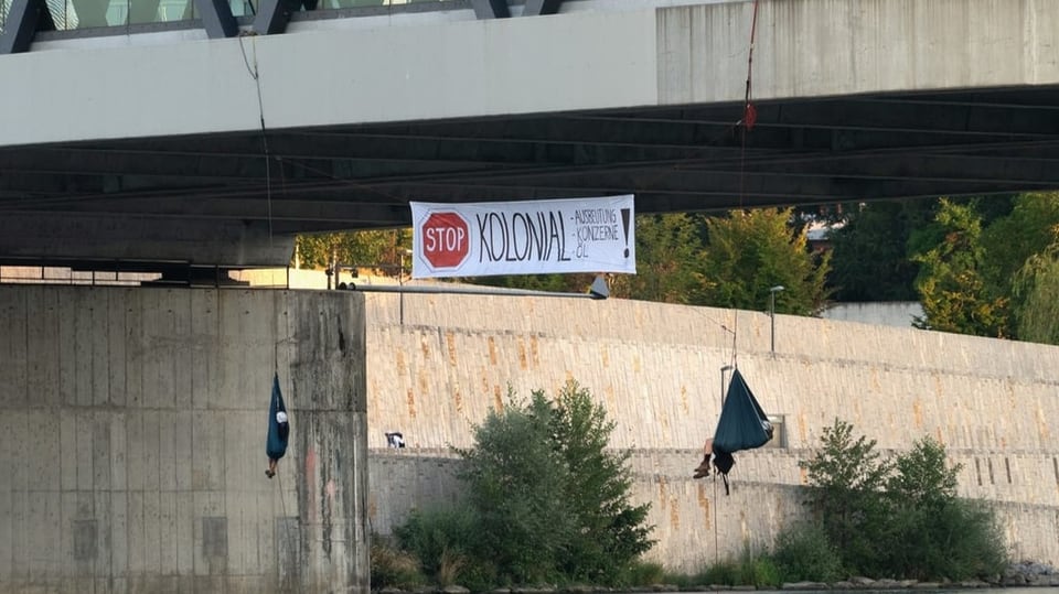 Ein Transparent mit der Aufschrift ' Stopp Kolonial' hängt von der Brücke, daneben hängen Menschen in Tüchern.