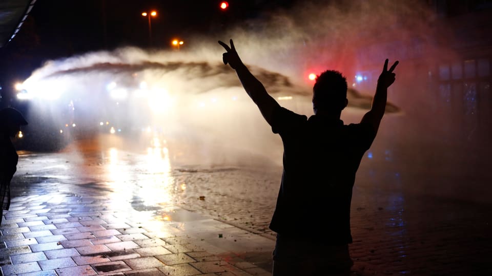 Ein junger Mann steht mit erhobenen Händen vor einem Wasserwerfer und macht das Peace-Zeichen.