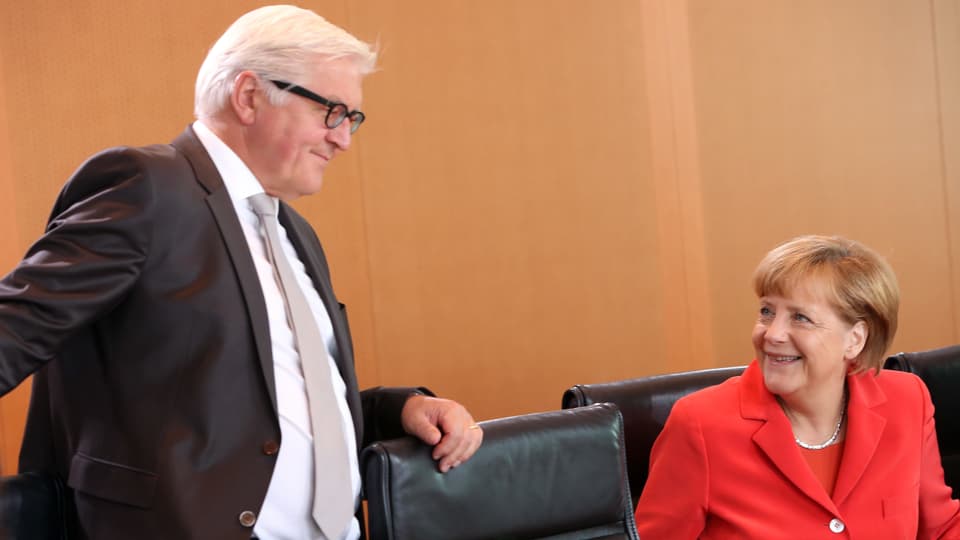 Steinmeier stehend und zur sitzenden Merkel hingewendet, die ihn anlächelt.